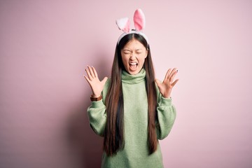 Sticker - Young asian woman wearing cute easter bunny ears over pink background celebrating mad and crazy for success with arms raised and closed eyes screaming excited. Winner concept