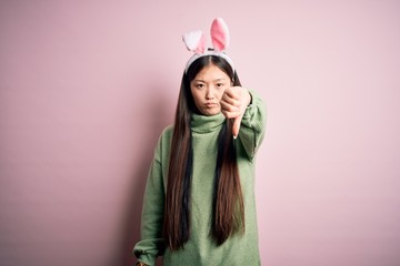 Canvas Print - Young asian woman wearing cute easter bunny ears over pink background looking unhappy and angry showing rejection and negative with thumbs down gesture. Bad expression.