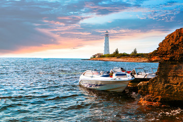 Two boats are moored at the rocky coast of the black sea. In the background, a coastal lighthouse. Cape Tarhankut. Horizontal photo. Summer. Evening. Sunset. Space for text