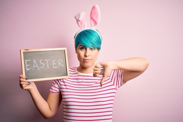 Sticker - Young woman with fashion blue hair wearing easter rabbit ears and holding blackboard with holiday word with angry face, negative sign showing dislike with thumbs down, rejection concept