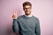 Young handsome man with beard wearing glasses and sweater standing over pink background showing and pointing up with fingers number two while smiling confident and happy.