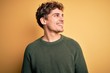 © Krakenimages.com - Young blond handsome man with curly hair wearing green sweater over yellow background looking away to side with smile on face, natural expression. Laughing confident.