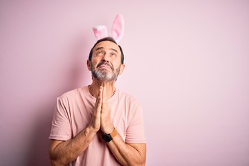 Sticker - Middle age hoary man wearing bunny ears standing over isolated pink background begging and praying with hands together with hope expression on face very emotional and worried. Begging.