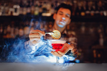Barman Is Preparing Red Alcoholic Cocktail With Blue Smoke In Martini Glass, Decorated With Orange