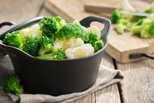 Black Saucepan With Cooked Broccoli And Cauliflower On Wooden Kitchen Table.