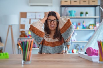 Young beautiful teacher woman wearing sweater and glasses sitting on desk at kindergarten suffering from headache desperate and stressed because pain and migraine. Hands on head.