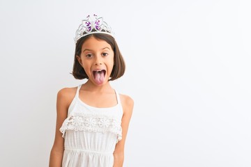 Beautiful child girl wearing princess crown standing over isolated white background sticking tongue out happy with funny expression. Emotion concept.