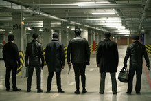 Rear View Of Row Of Criminals Or Gangsters In Black Standing On Parking Area