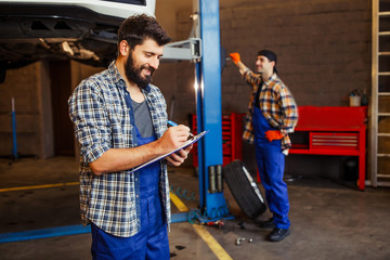 two automechanics working with lifted car in workshop, one checking car and another taking notes in clipboard