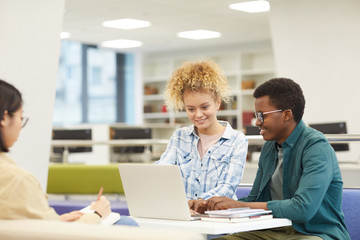 Wall Mural - Multi-ethnic group of students using laptop and smiling happily while working on project in college library, copy space