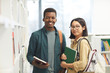 © Seventyfour - Waist up portrait of two international students looking at camera and smiling while posing by shelves in college library, copy space