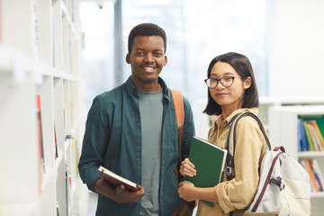Wall Mural - Waist up portrait of two international students looking at camera and smiling while posing by shelves in college library, copy space