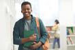 © Seventyfour - Waist up portrait of African-American student looking at camera and smiling while posing by shelves in college library, copy space
