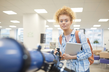 Wall Mural - Portrait of cheerful female student looking at camera while posing by telescope in astronomy class or college library, copy space