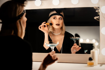 Poster - Beautiful blonde woman in twenties years clothes applying makeup near a mirror with bulbs