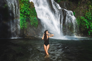 Wall Mural - Young slim brunette woman with curly hair enjoying in lagoon of huge tropical waterfall Banyumala in Bali. Wearing in black swimsuit. Happy vacations in Indonesia. Wanderlust travel concept.