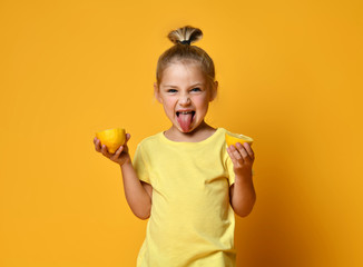 Wall Mural - Little smiling cute girl in yellow t-shirt holding halves of fresh sour ripe lemon fruit and showing tongue over yellow background