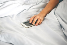 Close-up Hand Holds The Phone In Bed. The Girl Fell Asleep With A Phone In Her Hand And She Is Addicted. Or A Person Reaches For A Mobile Phone To Turn Off The Alarm.