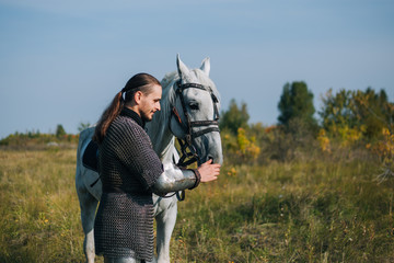 A knight strokes a white horse. Armored guy with a white horse. A man in chain mail. Image of a warrior