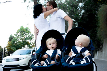 Wall Mural - happy parents kissed on the street and walking with twins sit on the buggy. The kid watching to the parent .