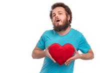 Happy Valentines Day. Crazy Bearded Man With Funny Curly Hair In Blue T-shirt. Silly Guy In Love, Isolated On White Background. Portrait Of Man With Red Heart Shaped Plush Pillow, Look At Camera