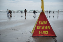A Red And Yellow Swim Between Flags Lifeguards Safety Warning Sign On Piha Beach On Auckland's West Coast.  Selective Focus With People Behind Walking On The Beach And Playing In The Sea And Waves.