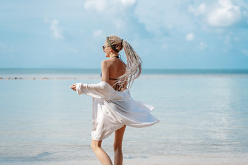 Wall Mural - very slender blonde with dreadlocks against a tropical landscape with a blue sea and sky and, in a white shirt and sunglasses, tropical vacation and travel concept