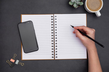 Blank open notebook with empty page, coffee cup and hand holding a pencil. Table top, work space ondark ,textured black background. Creative flat lay.