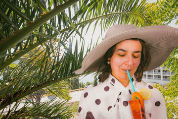 Summer day, vacation trip. Beautiful woman in big hat drinking a fresh cocktail under a palm tree.