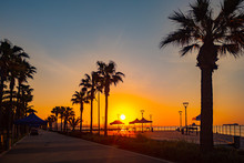 Cyprus. Limassol Promenade At Sunset. Sunset On The Mediterranean Sea. The Molos Waterfront In Limassol. Silhouettes Of Palm Trees, Lanterns And Umbrellas Against The Setting Sun. Holidays In Cyprus.