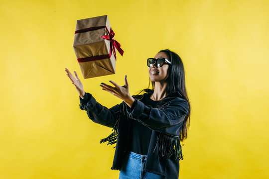Portrait of a happy woman throws up a gift box and looking at camera over yellow background
