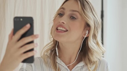 Poster - An attractive smiling woman with blond hair is having a video call on her cellphone with wired headphones isolated over fabric background