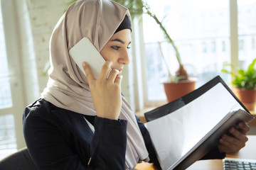 Wall Mural - Talking on phone while reviewing docs. Beautiful arabian businesswoman wearing hijab while working at openspace or office. Concept of occupation, freedom in business area, success, modern solution.