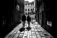Grayscale Shot Of Two Men Walking Down An Alley Casting Shadows On The Foreground