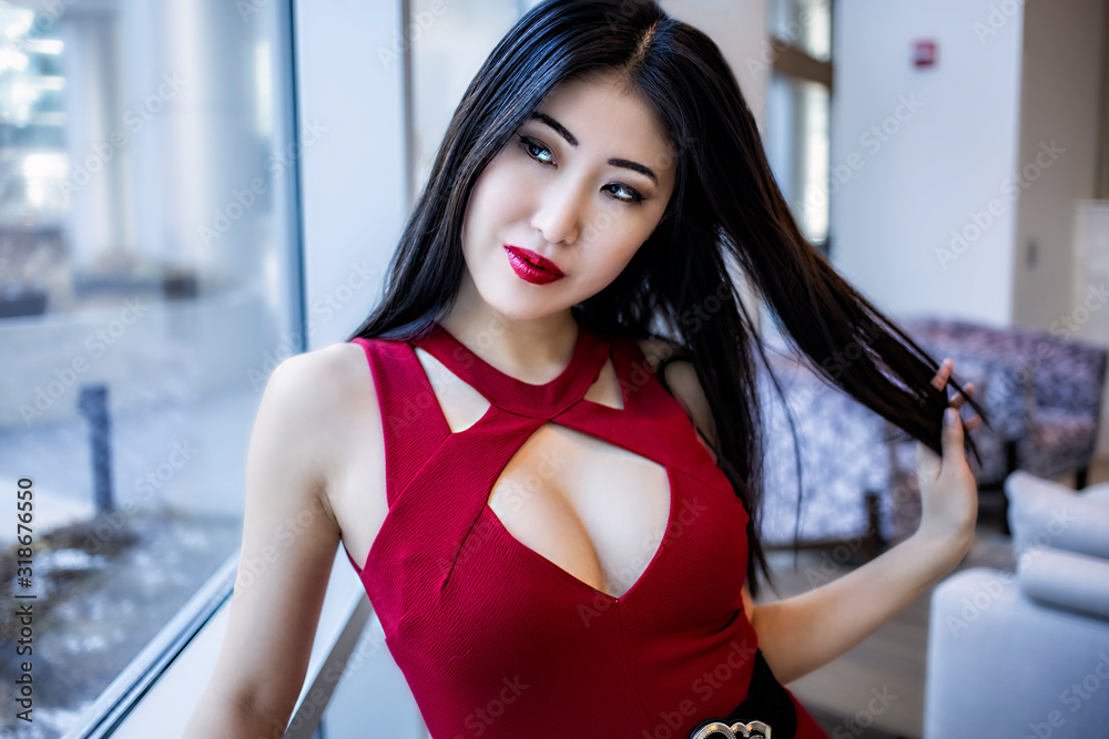 Fotografia do Stock beautiful female Asian model wearing a fashionable  elegant red dress and glossy red lipstick. posing sensually in a luxury  condo downtown. Chinese woman has long black hair and gorgeous