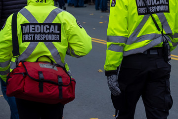 Two men dressed in bright reflective yellow jackets with medical first responder and a large x in reflective print on the backs of their coats. There's a red first aid bag on the attendant's shoulder.