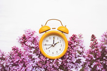 Yellow Alarm Clock And Bouquet Of Pink Lilacs On A White Surface, Top View. Hello Spring, Spring Time, Conceptual Image
