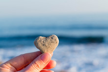Female Hand Holds A Heart Of Natural Stone Against The Background Of The Sea In The Sun. Reminder Of Mother's Day, Valentine's Day, Stone Wedding, Etc.