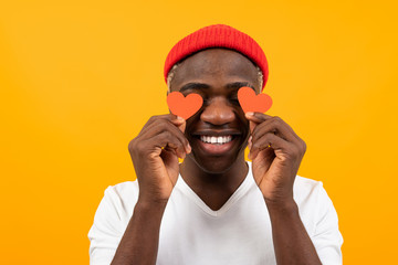 portrait of a handsome smiling american dark-skinned man in a white t-shirt holding two small eye-shaped postcards for Valentine's Day on a yellow background