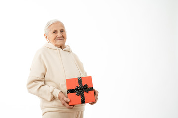 Confident old woman in beige hoodie holds red gift box and looking at the camera isolated over white background, copyspace for your text