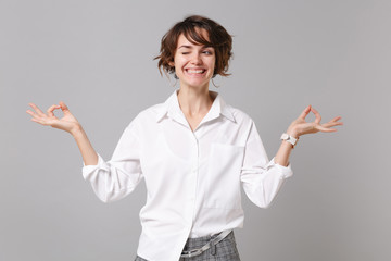 Wall Mural - Funny young business woman in white shirt posing isolated on grey background. Achievement career wealth business concept. Mock up copy space. Hold hands in yoga gesture, relaxing meditating, blinking.