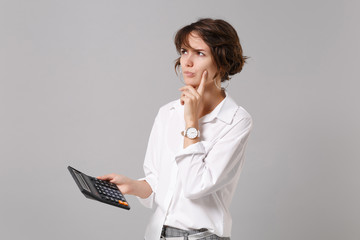 Sticker - Pensive young business woman in white shirt posing isolated on grey wall background in studio. Achievement career wealth business concept. Mock up copy space. Hold calculator put hand prop up on chin.