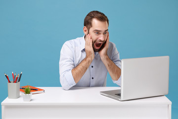 Wall Mural - Surprised young man in light shirt sit at desk isolated on pastel blue background. Achievement business career concept. Mock up copy space. Work on project with laptop pc computer put hands on cheeks.