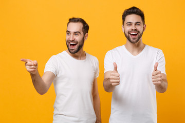 Sticker - Funny young men guys friends in white blank empty t-shirts posing isolated on yellow orange background. People lifestyle concept. Mock up copy space. Showing thumbs up, pointing index finger aside.