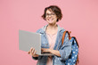 © ViDi Studio - Cheerful young woman student in denim clothes glasses backpack isolated on pastel pink background. Education in high school university college concept. Mock up copy space. Work on laptop pc computer.