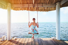 A Woman Sitting On A Swing On A Dock Overlooking The Ocean