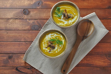 Pumpkin cream soup with cream and mushrooms in two wooden bowls on wooden background