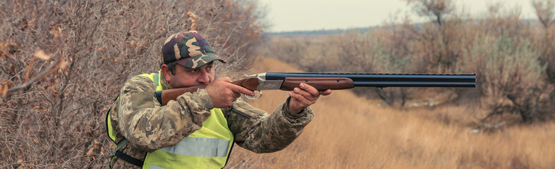 A man with a gun in his hands and an green vest on a pheasant hunt in a wooded area in cloudy weather. Hunter with dogs in search of game.