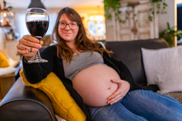 A shallow depth of field as a blurry pregnant woman relaxes on sofa in family room with glass of red wine, risking fetal alcohol syndrome. Copy space to right