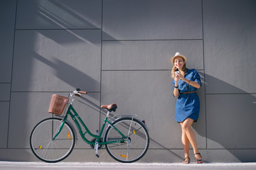 Wall Mural - Lifestyle and technology. Young pretty woman using smartphone with bicycle leaning on the grey wall.
